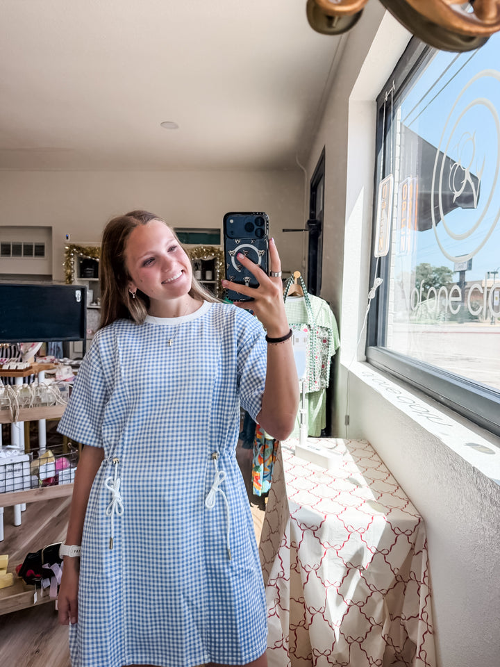 A woman taking a mirror selfie in a boutique shop, wearing a light blue and white gingham short-sleeve dress with drawstrings at the waist.