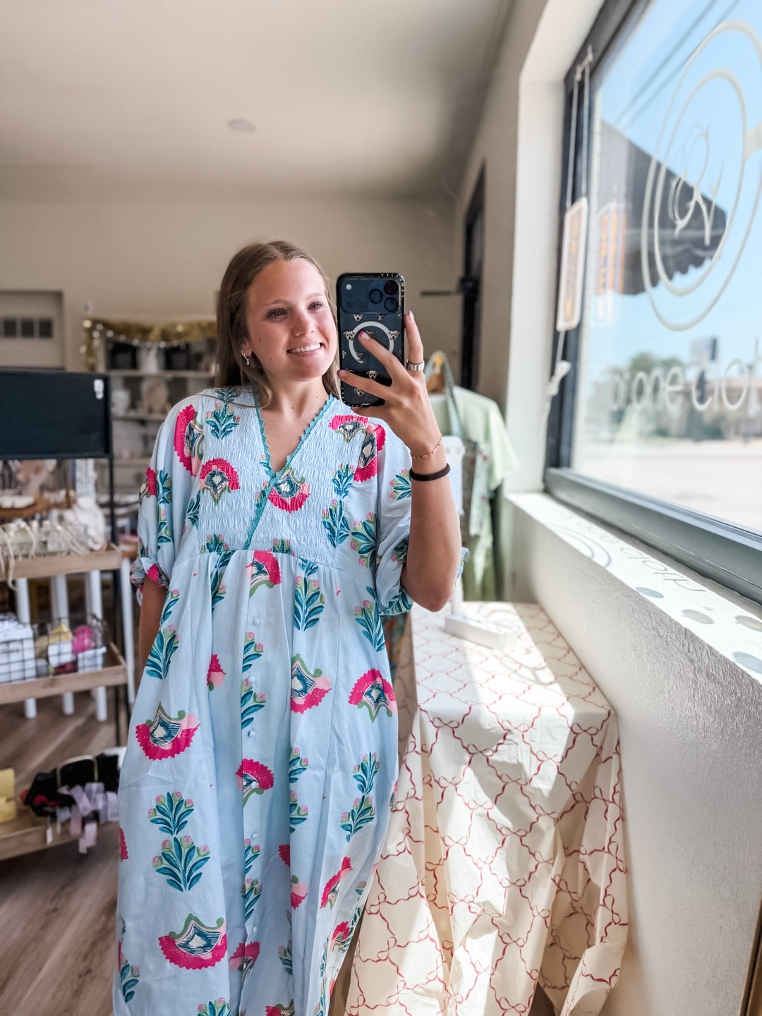 A woman takes a mirror selfie wearing a light blue kaftan dress with a pink and green tropical floral print in a boutique setting.