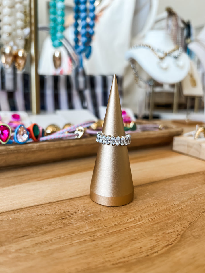 A silver-toned diamond band ring displayed on a metallic gold cone jewelry stand, set on a wooden table with a blurred boutique background.