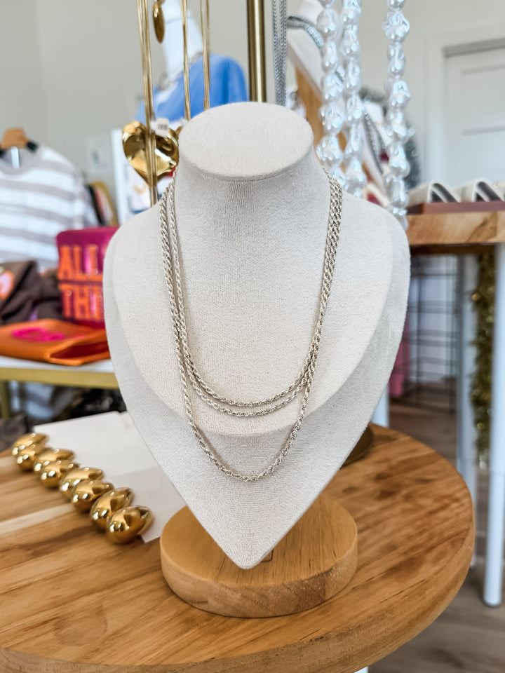 Three strands of silver rope chain necklaces displayed on a white fabric jewelry bust, set on a round wooden table in a boutique setting.
