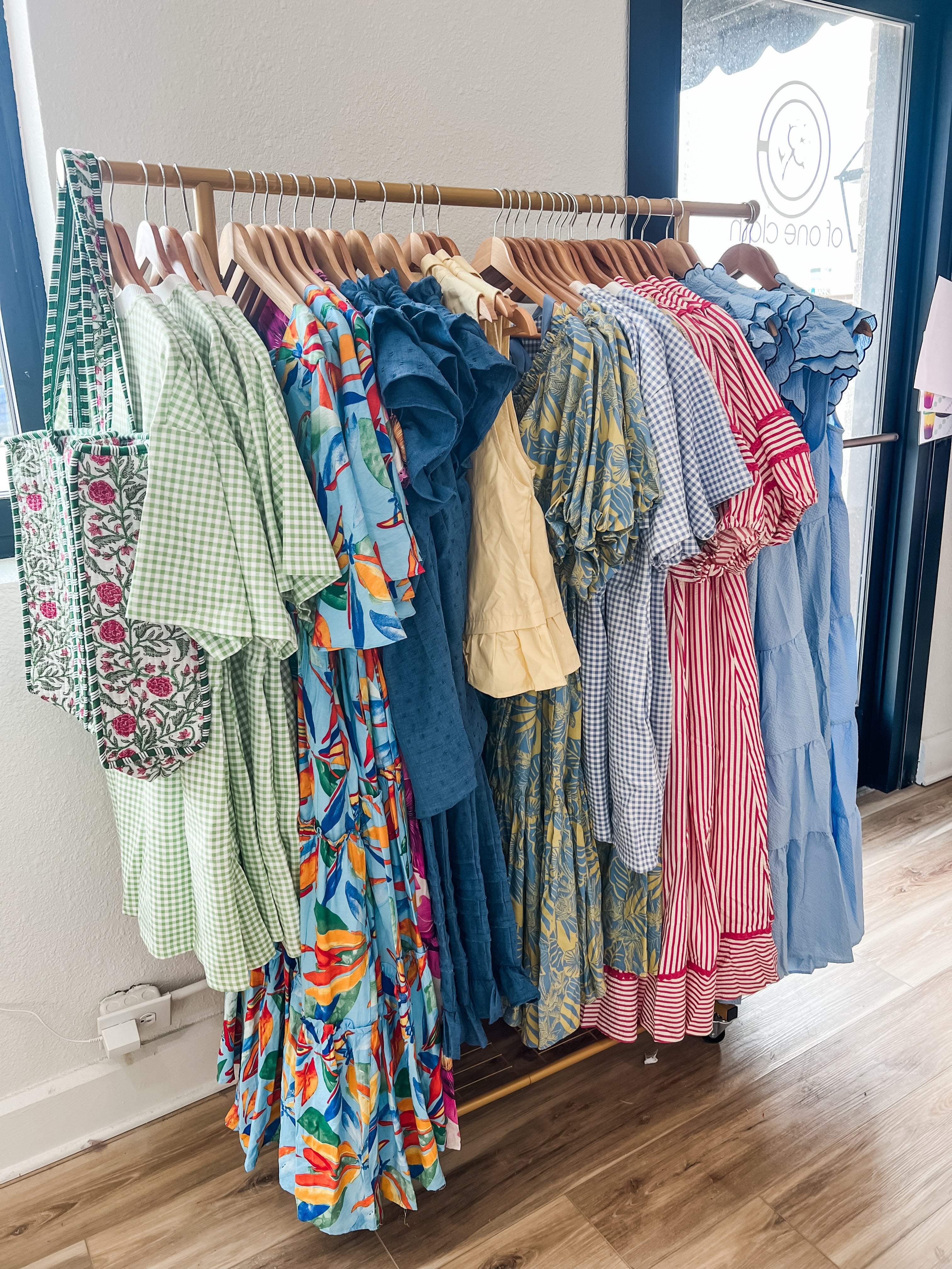 A clothing rack filled with multiple colorful dresses and tops in various patterns including gingham, floral, and stripes, set in a boutique with wood floors and a window.