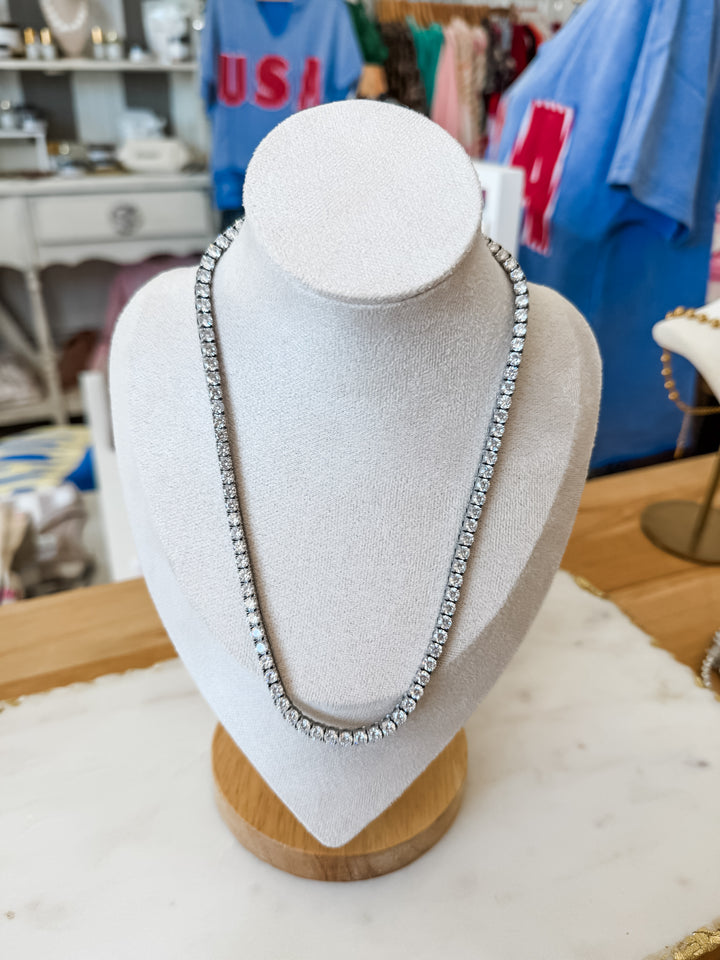 A silver tennis necklace displayed on a white fabric jewelry bust with a wooden base, set in a retail store environment.