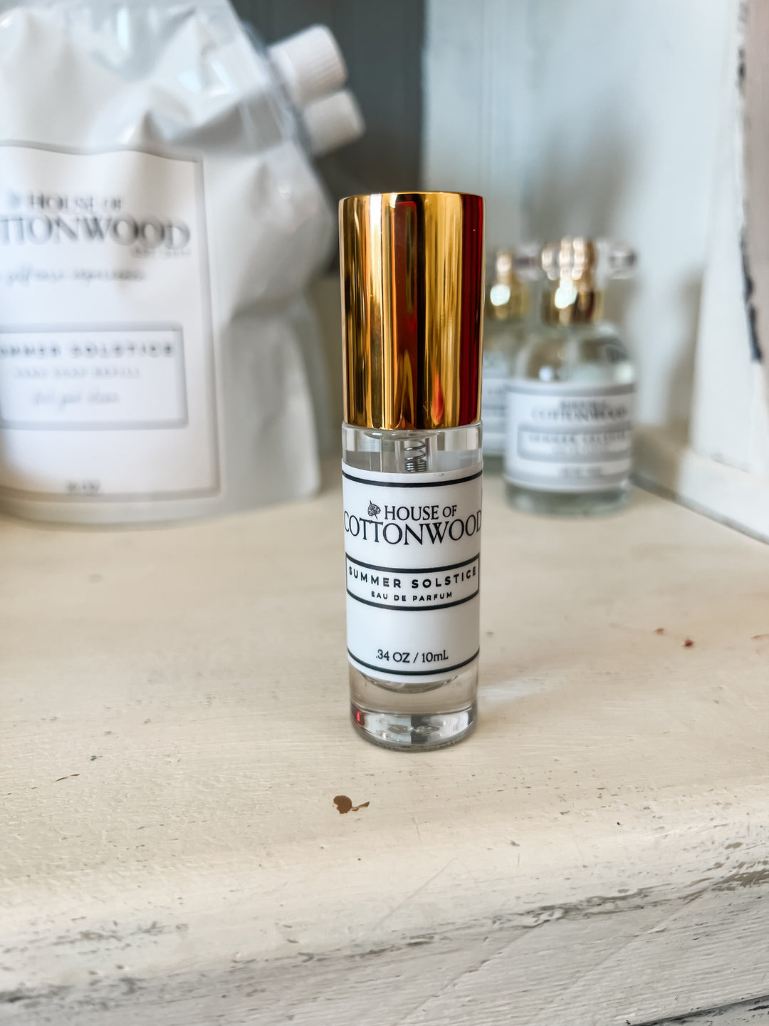 A 10mL spray bottle of House of Cottonwood Summer Solstice Eau de Parfum standing on a light wooden surface, with its white and gold cap facing forward. The background shows other House of Cottonwood fragrance bottles and a white bag in a softly blurred setting.