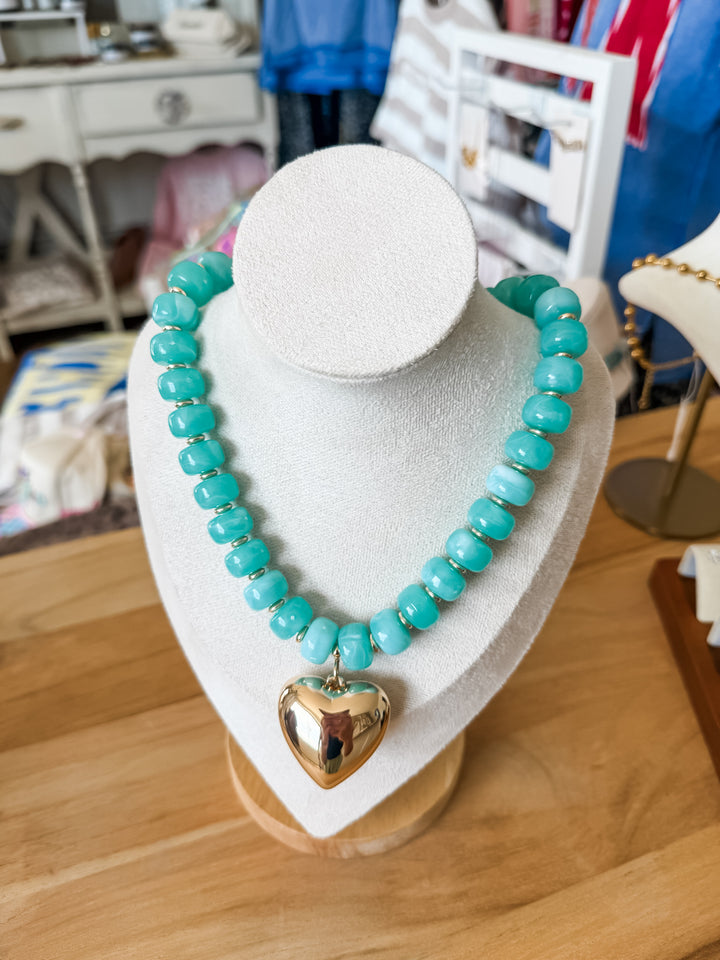 A turquoise beaded necklace with a polished gold heart pendant, displayed on a white jewelry bust on a wooden table.