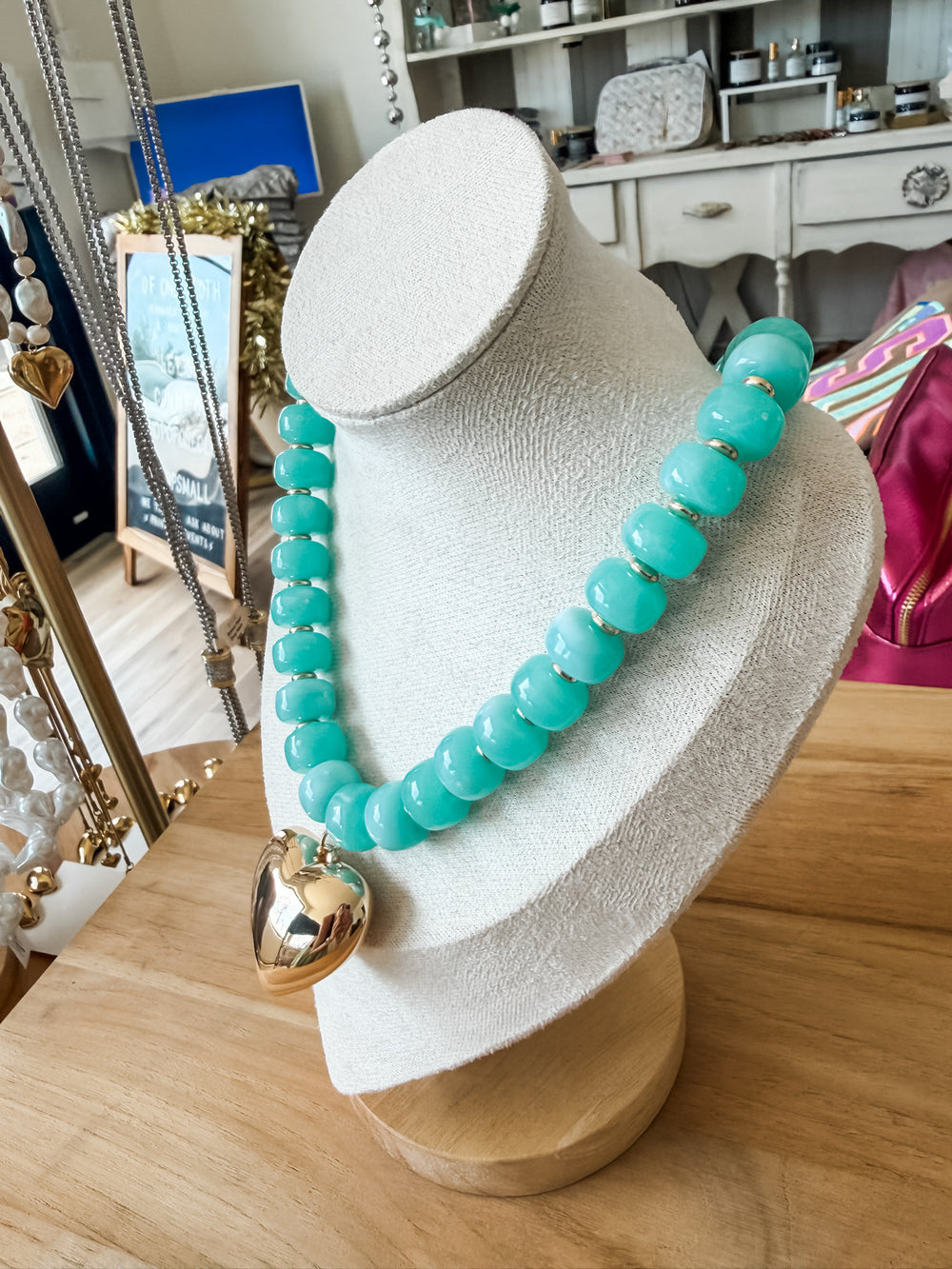 A turquoise beaded necklace with a gold heart pendant displayed on a white jewelry bust in a boutique setting.