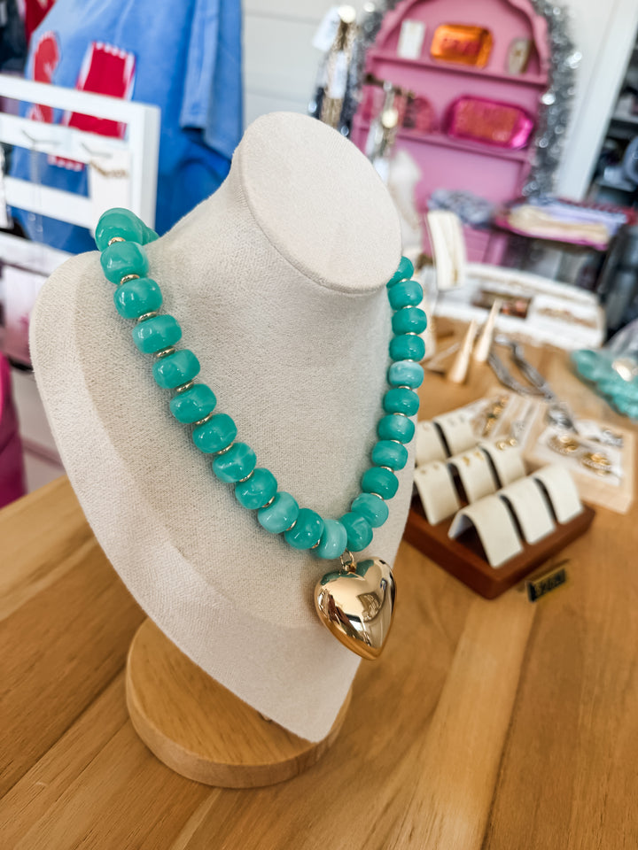 A turquoise beaded necklace with a gold heart pendant displayed on a white jewelry bust in a retail setting.