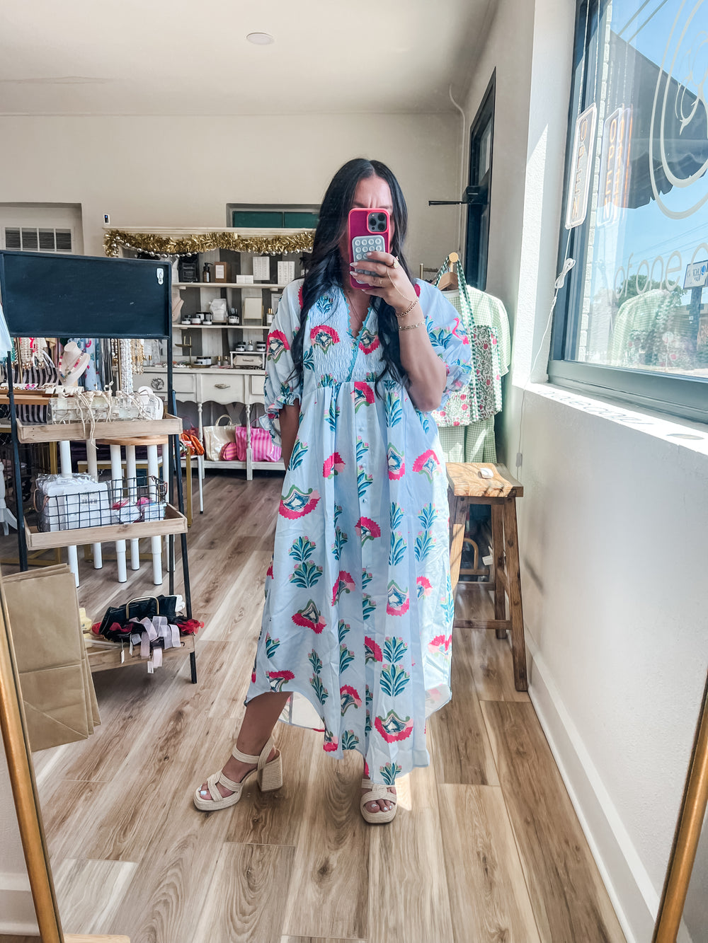 A woman takes a mirror selfie in a boutique wearing a light blue maxi dress with a pink floral and green tree pattern. The boutique has wood floors, shelves, and clothing racks in the background.