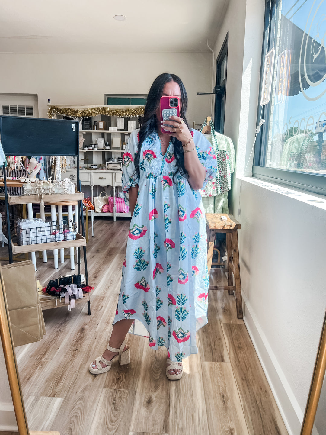 A woman takes a mirror selfie in a boutique wearing a light blue maxi dress with a pink floral and green tree pattern. The boutique has wood floors, shelves, and clothing racks in the background.