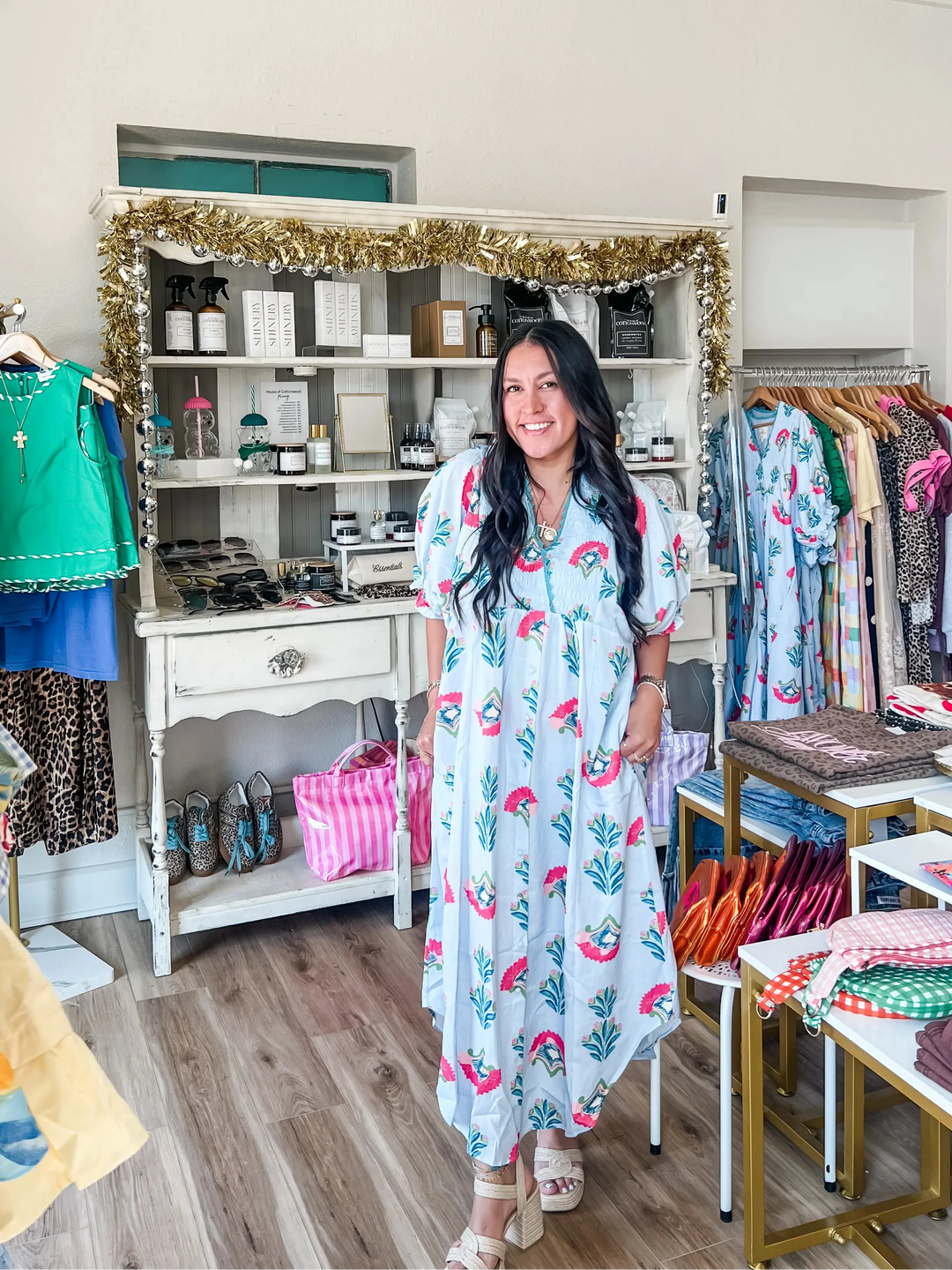 A woman stands in a boutique wearing a light blue maxi dress with a pink floral print. The shop is filled with clothing racks, shelves, and display tables.