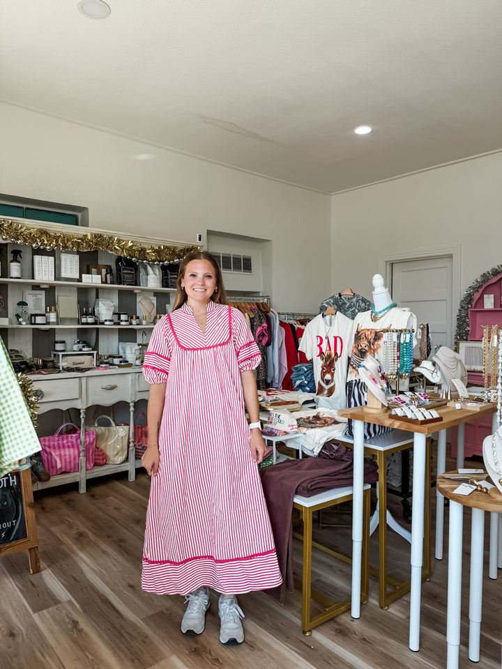 A woman wearing a pink and white vertically striped short-sleeve dress stands in a retail boutique. The room has wood floors, white walls, shelves with various items, and clothing displays in the background.