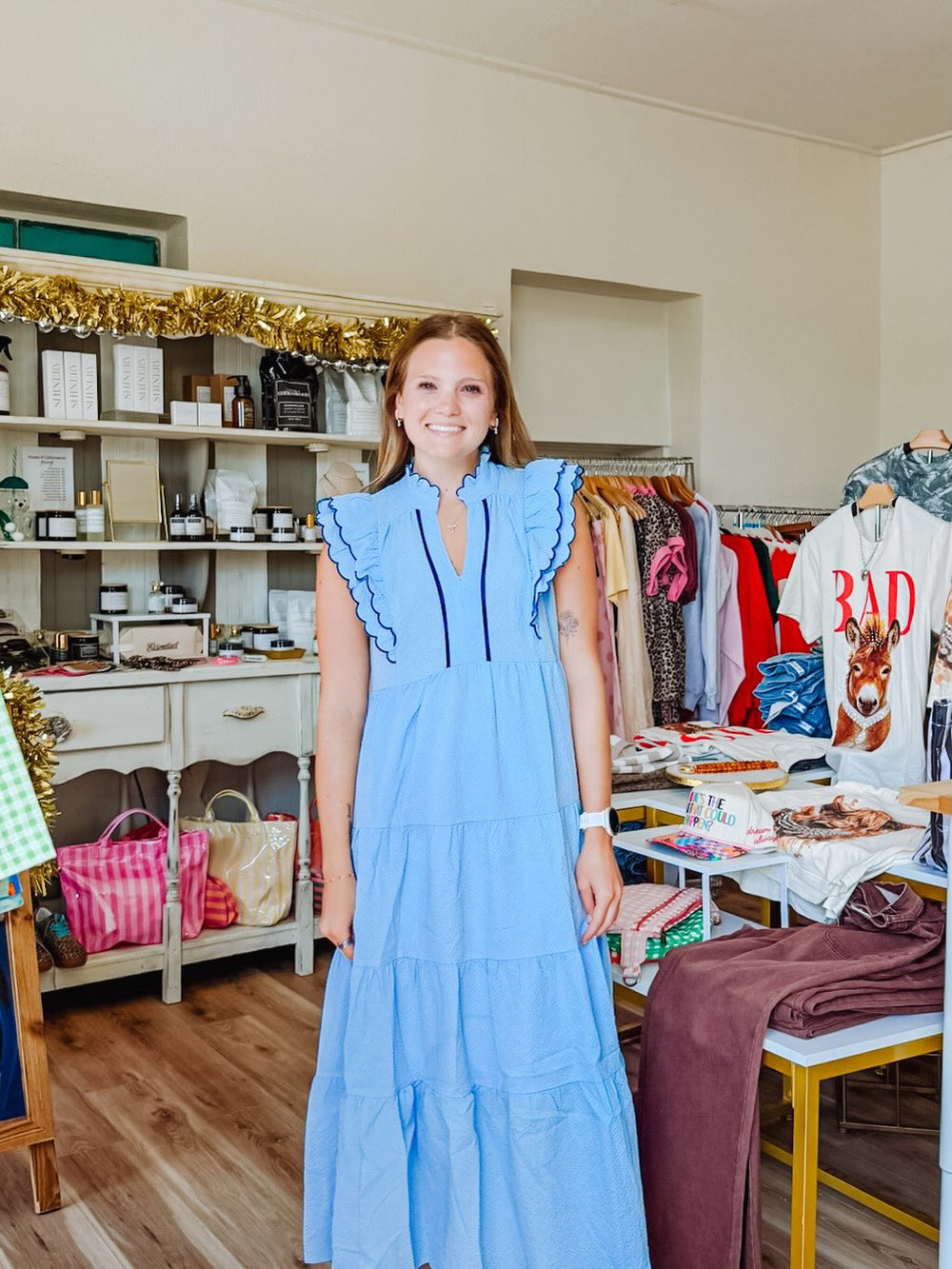 A woman wearing a light blue tiered maxi dress with ruffle details on the shoulders and black piping along the neckline, standing in a boutique interior with clothing racks and shelves visible in the background.