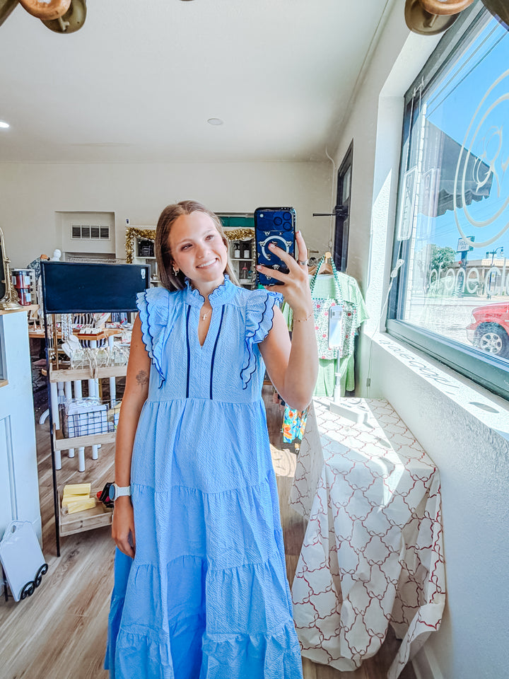 A woman takes a mirror selfie in a boutique while wearing a light blue tiered maxi dress with ruffled shoulder details. The shop interior is visible, including a window, patterned table runner, and clothing racks.