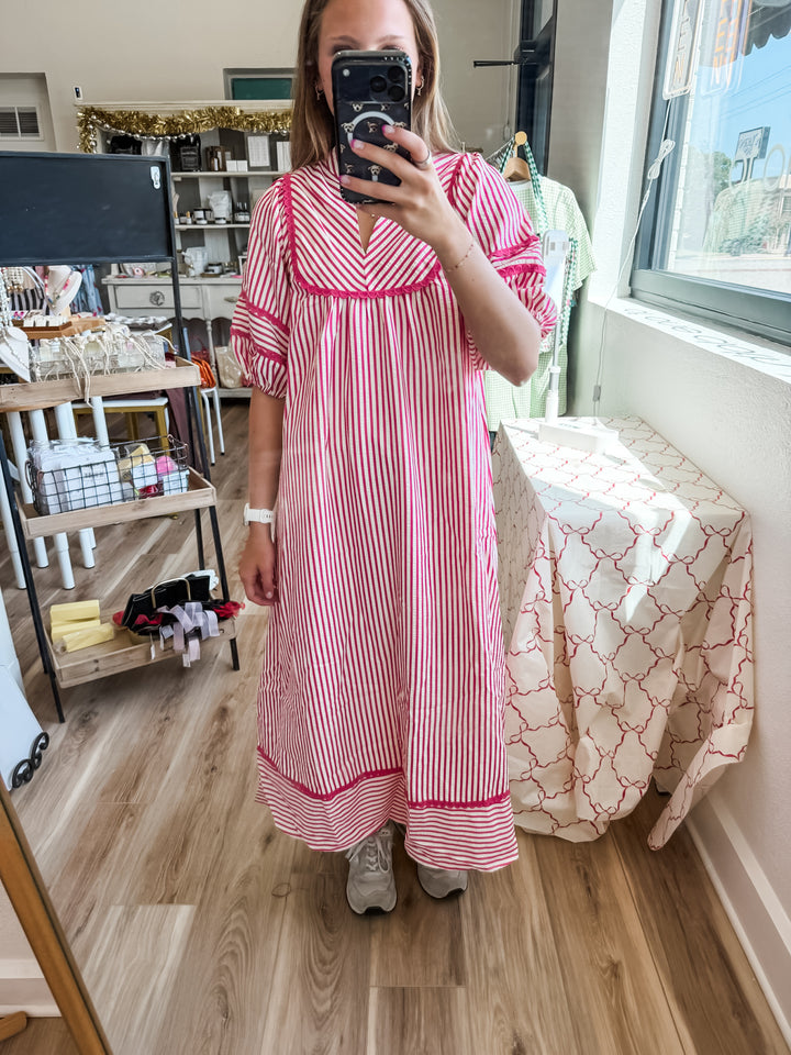 A woman takes a mirror selfie wearing a pink and white vertically striped midi dress with a V-neck and short puff sleeves, standing in a retail shop.