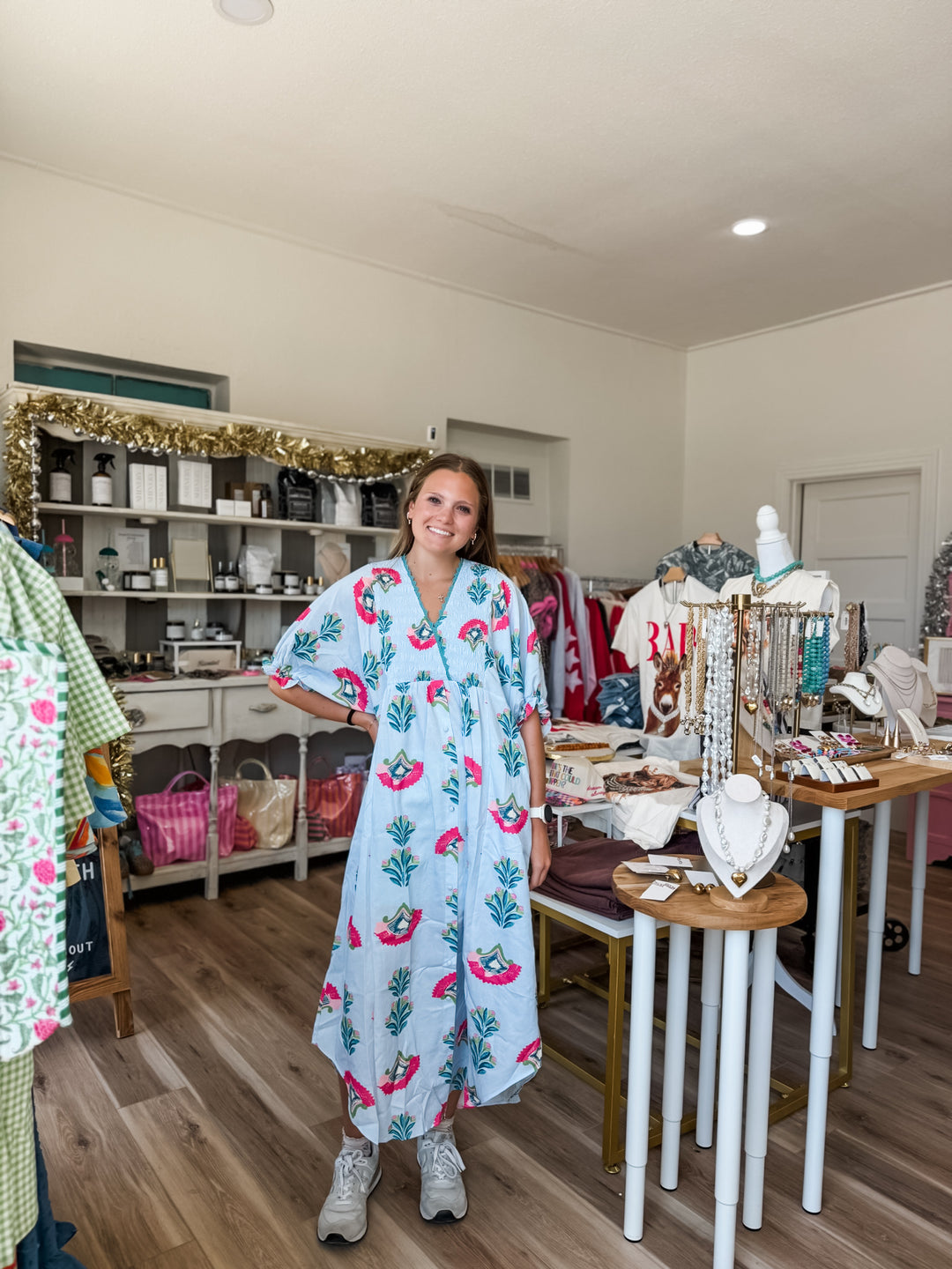 A woman stands in a boutique wearing a light blue kaftan dress with a pink flamingo and green palm tree pattern. The shop interior features wooden floors, display tables with jewelry, and shelves in the background.