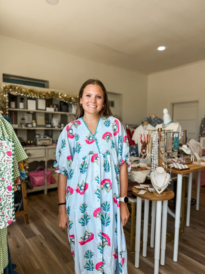 A woman stands in a boutique wearing a light blue kaftan dress with a pink and green floral print. The shop interior features wooden floors, display tables with jewelry, and shelves in the background.