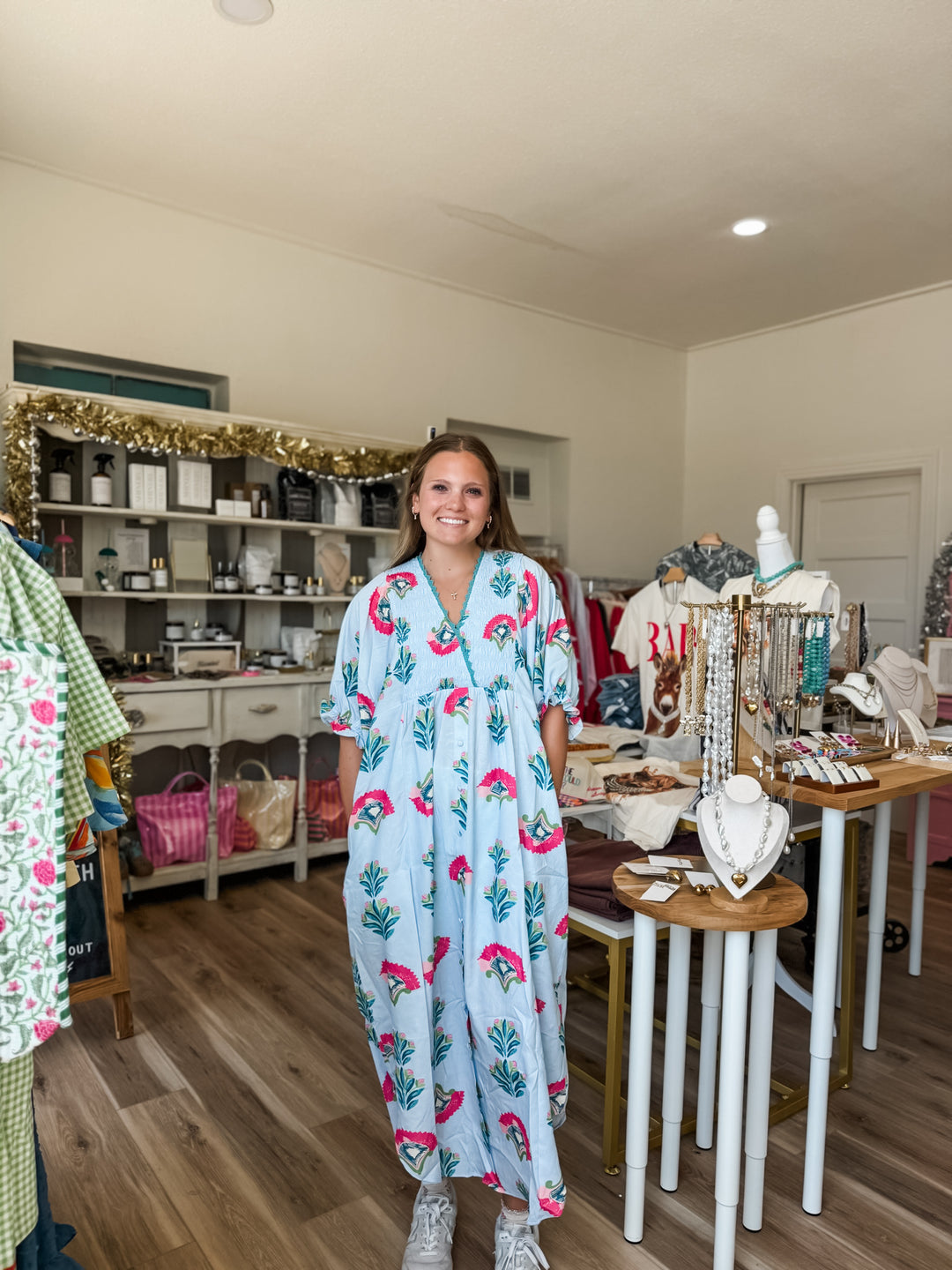 A woman stands in a boutique wearing a light blue kaftan dress with a pink floral and green leaf pattern. The shop interior features wooden floors, display tables with jewelry, and shelves in the background.