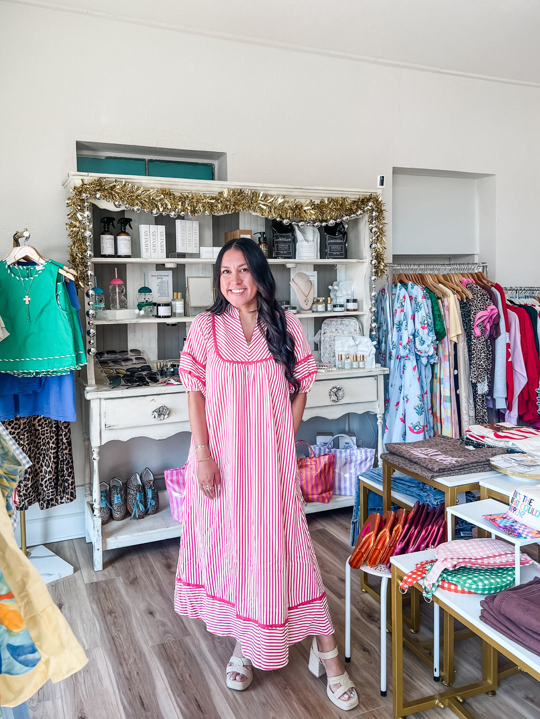 A woman wearing a pink and white vertically striped nightgown stands inside a boutique clothing store. The store features a white display cabinet with gold tinsel garland, racks of colorful clothing, and a wooden floor.