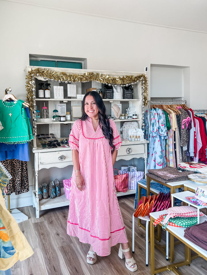 A woman wearing a pink and white vertically striped nightgown stands inside a boutique clothing store. The store features a white display cabinet with gold tinsel garland, racks of colorful clothing, and a wooden floor.