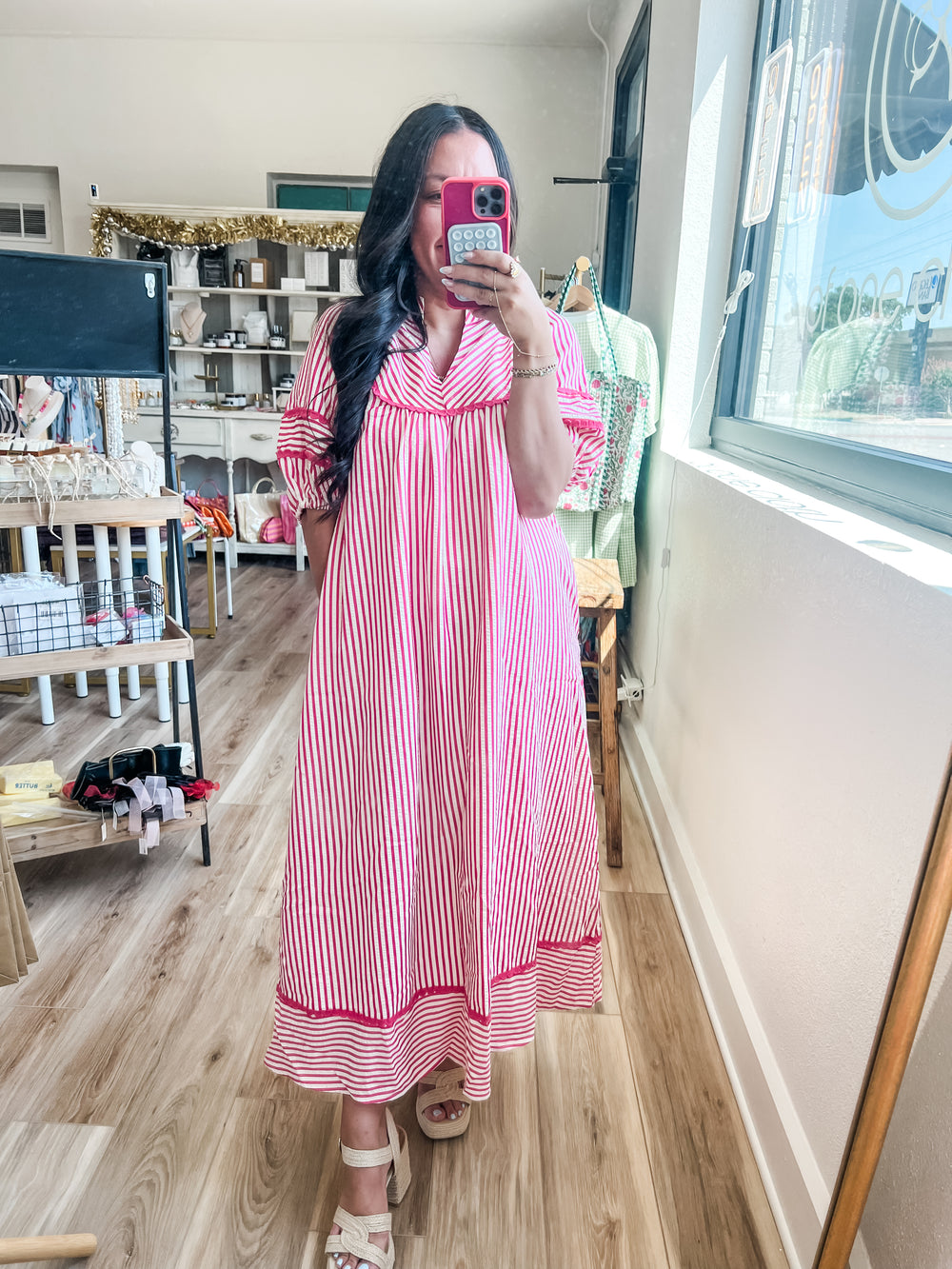 A woman takes a mirror selfie in a boutique while wearing a pink and white vertically striped maxi dress with a V-neck and ruffled sleeves. The boutique interior features light wood floors, shelves, and a window.