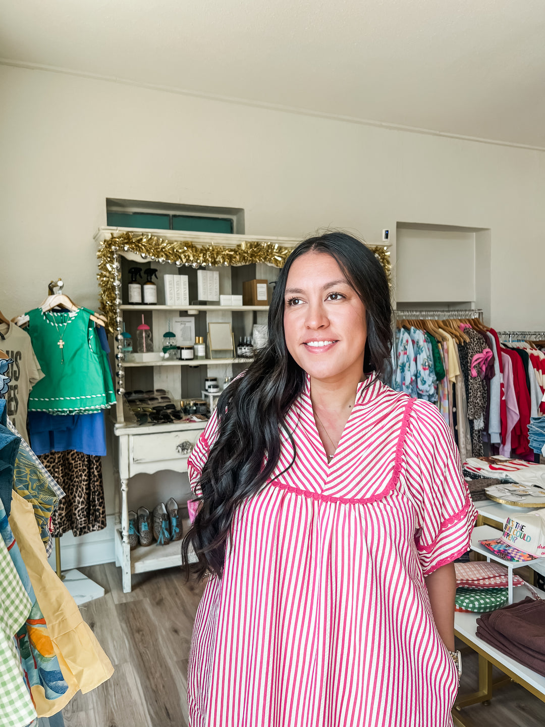A woman with long dark hair stands in a boutique wearing a pink and white vertically striped short-sleeve dress with pink trim. The background shows clothing racks, shelves, and a display cabinet inside the shop.