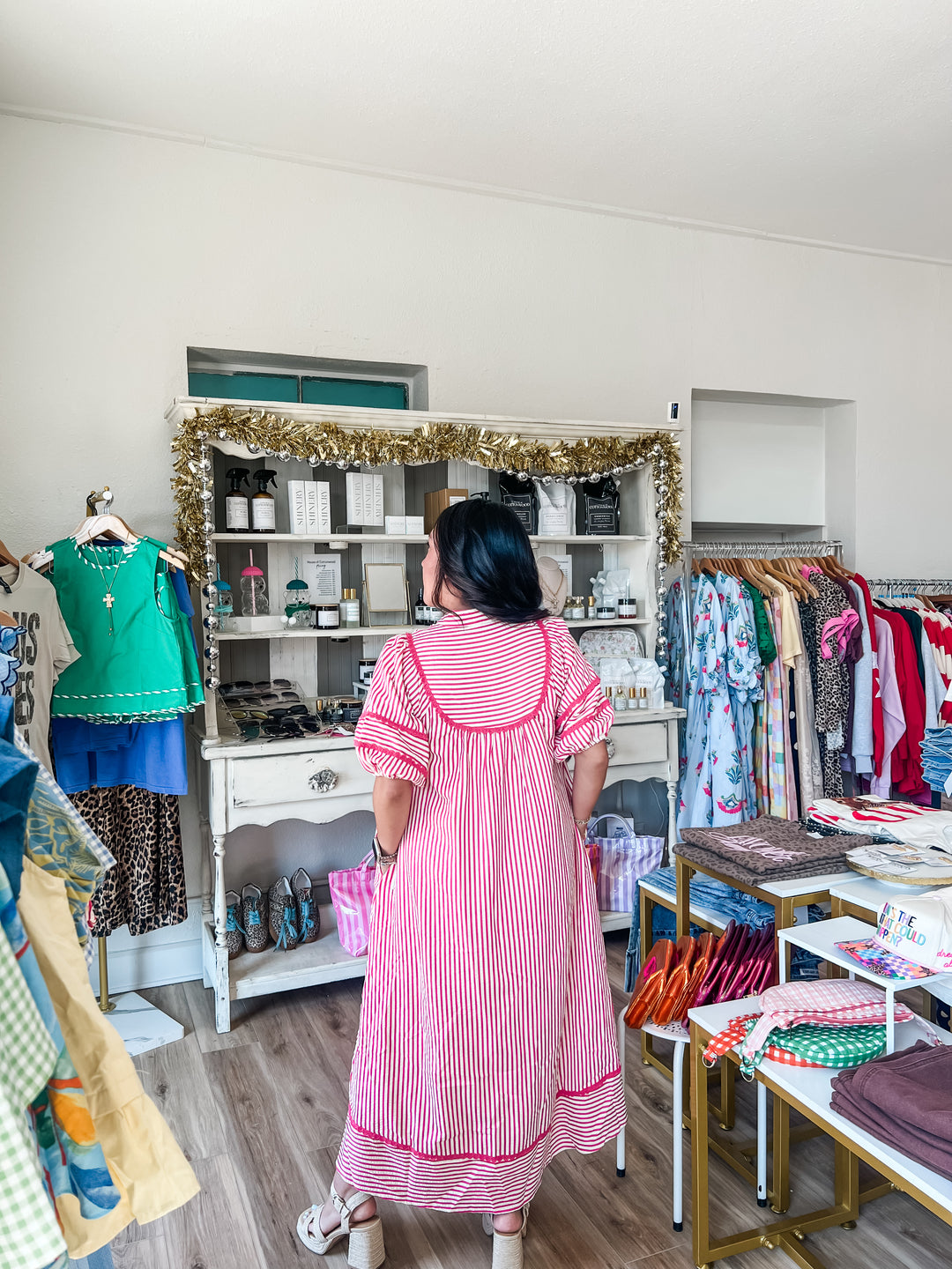 A woman viewed from behind wearing a pink and white vertically striped dress with red trim, standing inside a boutique clothing store with racks of garments, a white display cabinet, and wood flooring.