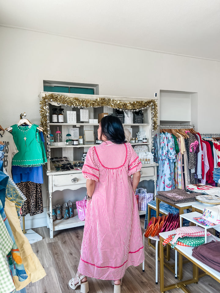 A woman viewed from behind wearing a pink and white vertically striped dress with red trim, standing inside a boutique clothing store with racks of garments, a white display cabinet, and wood flooring.