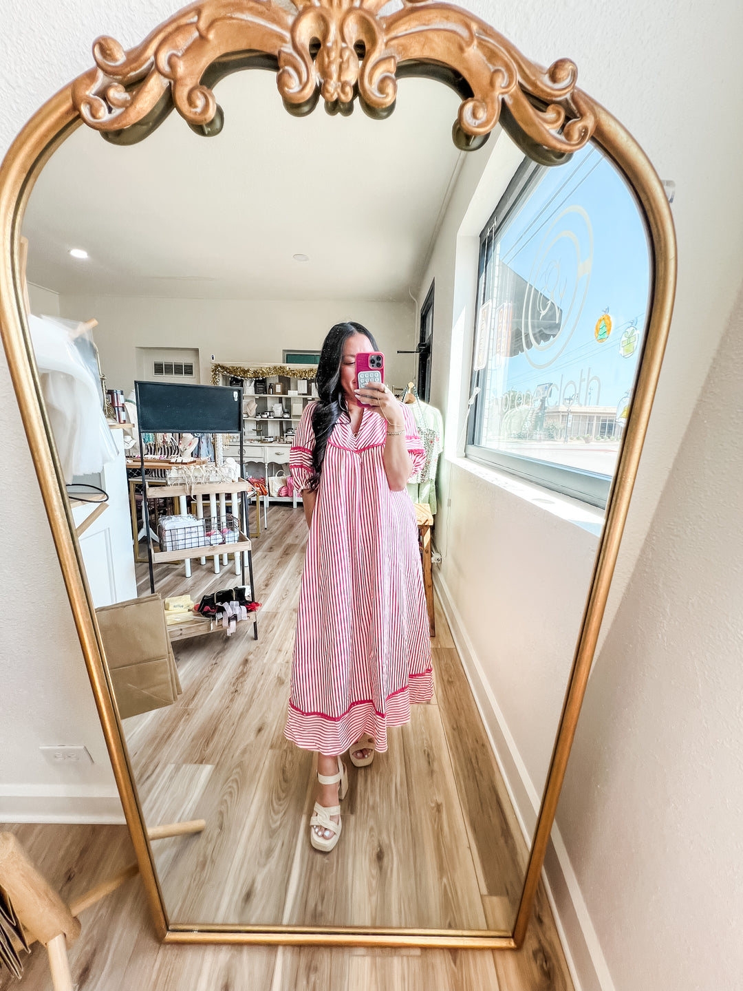 A woman takes a mirror selfie in a boutique, wearing a pink and white striped long-sleeve nightgown with a matching headband and white slide sandals. The background shows a shop interior with wooden floors, shelves, and a large ornate gold-framed mirror.