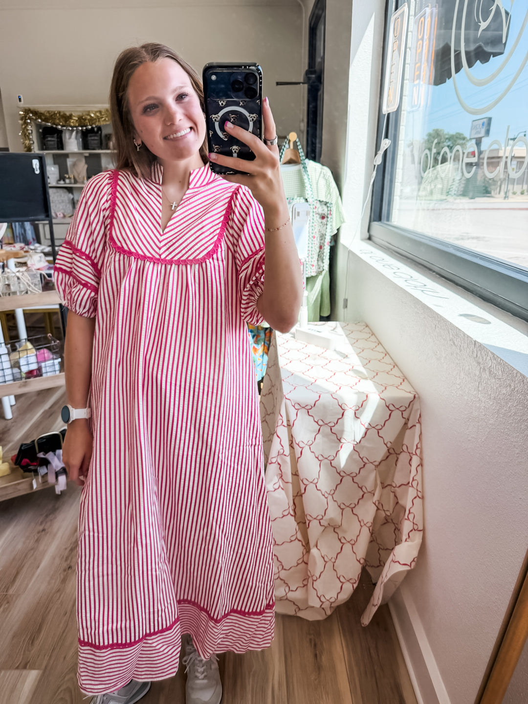 A woman takes a mirror selfie in a boutique wearing a pink and white vertically striped dress with bright pink trim. The background shows a retail shop interior with clothing racks, shelves, and a window.