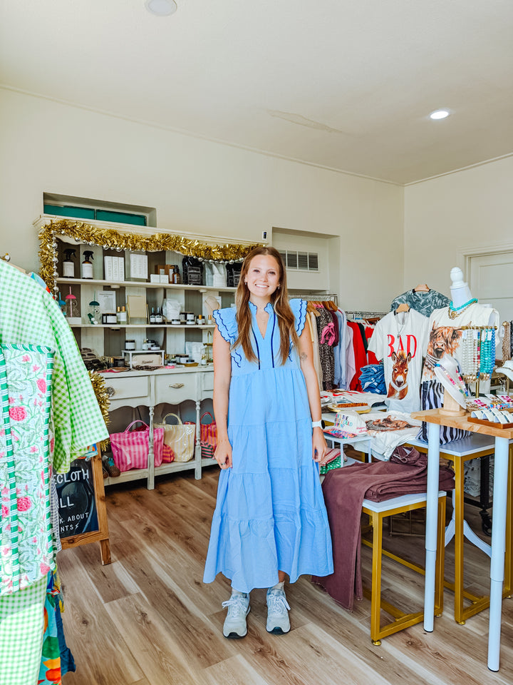 A woman stands in a boutique shop wearing a light blue tiered maxi dress. The shop has wood floors, white walls, shelves with various items, and clothing displays on tables and racks.