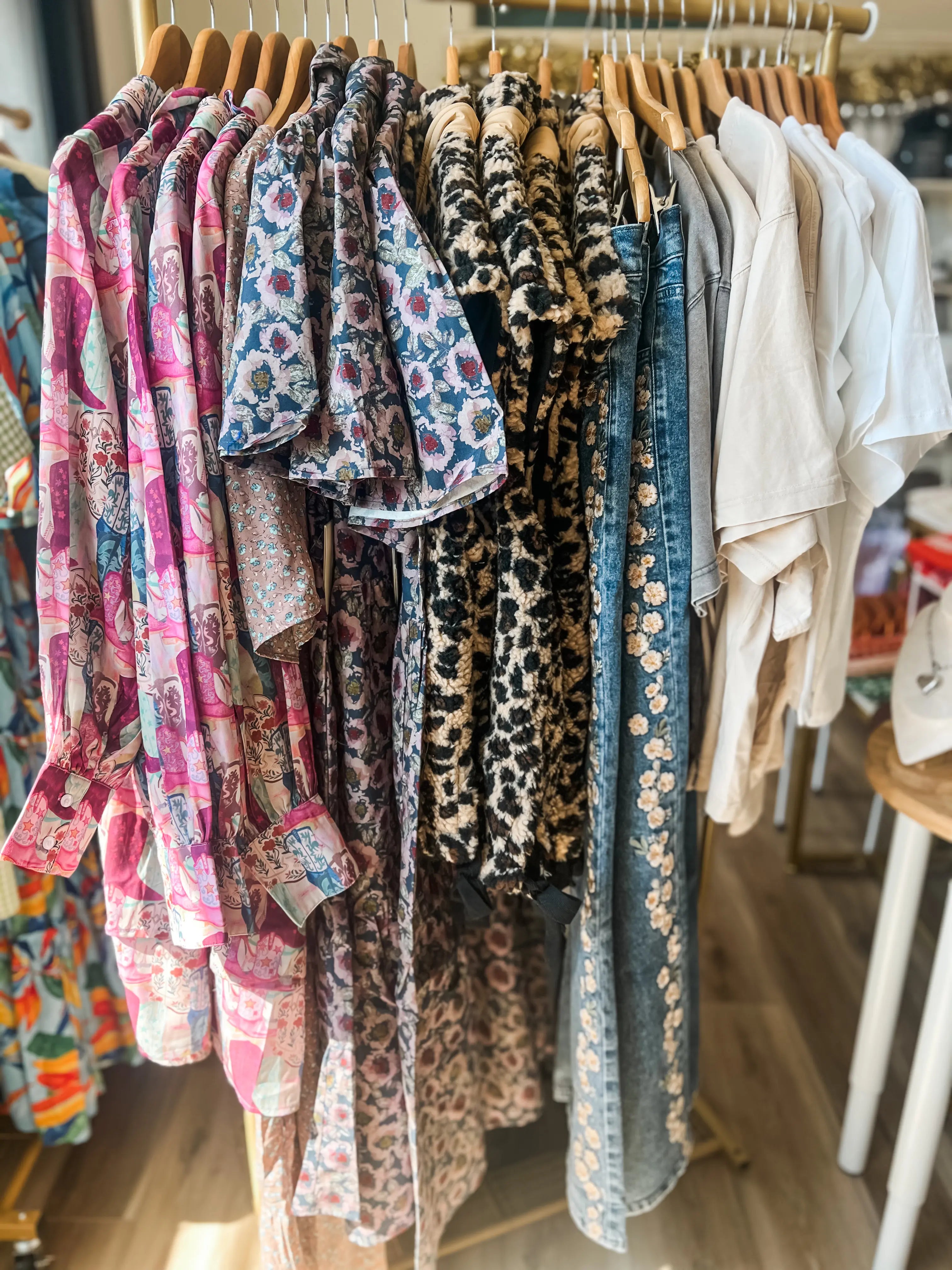 A clothing rack filled with multiple patterned and western solid shirts and dresses in a boutique setting. The garments include pink, blue, leopard print, and denim styles, hanging on wooden hangers.