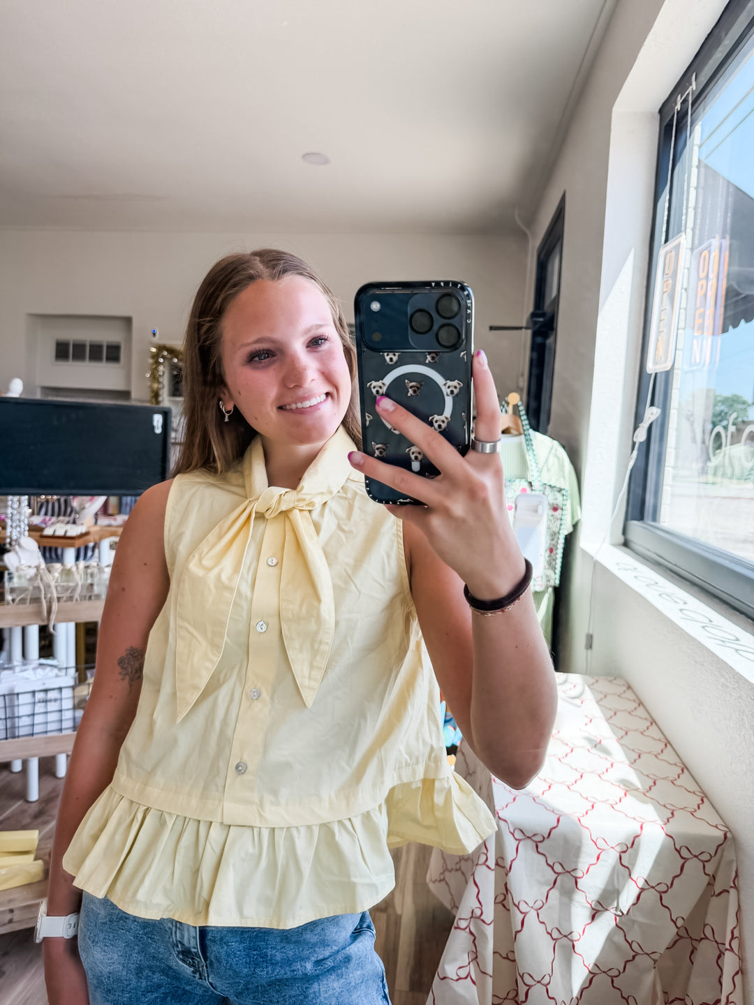 A woman taking a mirror selfie in a boutique, wearing a pale yellow sleeveless ruffled blouse with a large bow tie-neck and blue jeans.