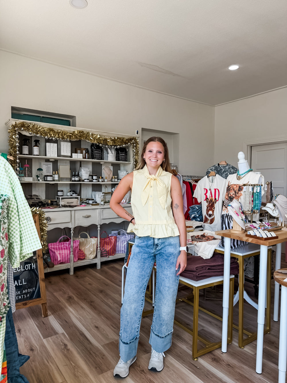 A woman stands in a boutique interior wearing a pale yellow sleeveless ruffled blouse and light-wash blue jeans. Shelves, tables, clothing racks, and store fixtures are visible around her.