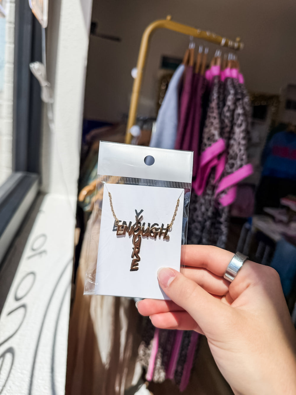 A hand holding a clear plastic card displaying a gold cross necklace with the "YOURE" vertically and the word 'ENOUGH' horizontally cut out to resemble a cross, with a clothing rack visible in the blurred background.