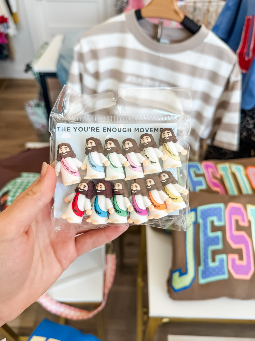 A hand holds a clear plastic bag containing 12 small Jesus figurines with the text 'THE YOU'RE ENOUGH MOVEMENT' printed on the packaging. The background shows a clothing store display with a striped shirt and a colorful pillow.