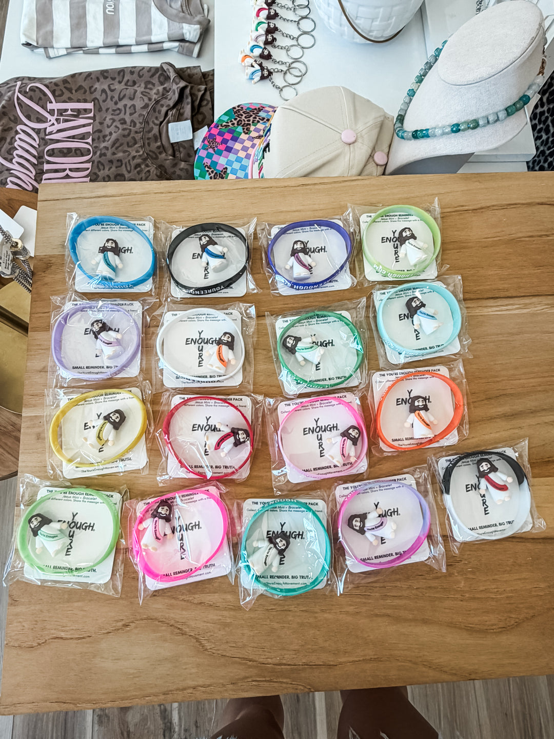 A set of 15 small packaged mini Jesus figurines + bracelet combos on white paper labels, arranged on a wooden tabletop. They come in multiple colors including blue, black, purple, green, yellow, orange, pink, and teal.