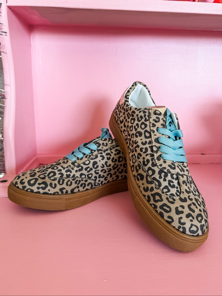 A pair of low-top sneakers with a beige and black leopard print pattern, bright turquoise blue laces, and brown gum soles, displayed on a pink shelf.