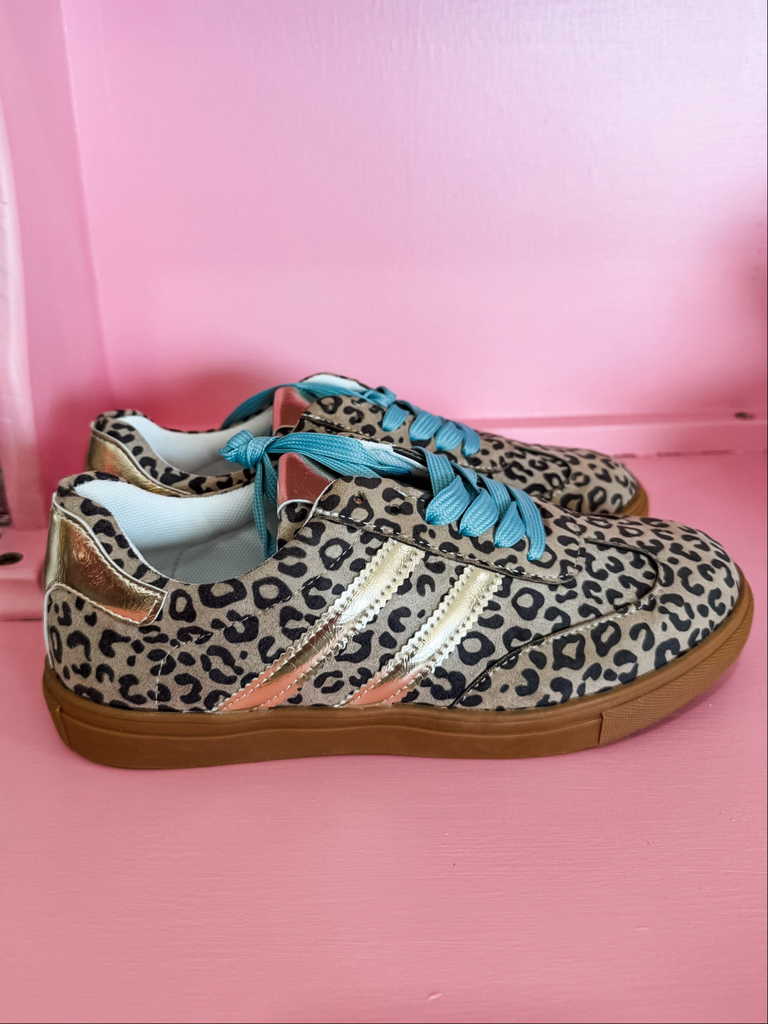 A pair of low-top sneakers with a beige, black, and brown leopard print pattern, featuring metallic gold side stripes, turquoise blue laces, and brown gum soles, displayed on a pink surface.