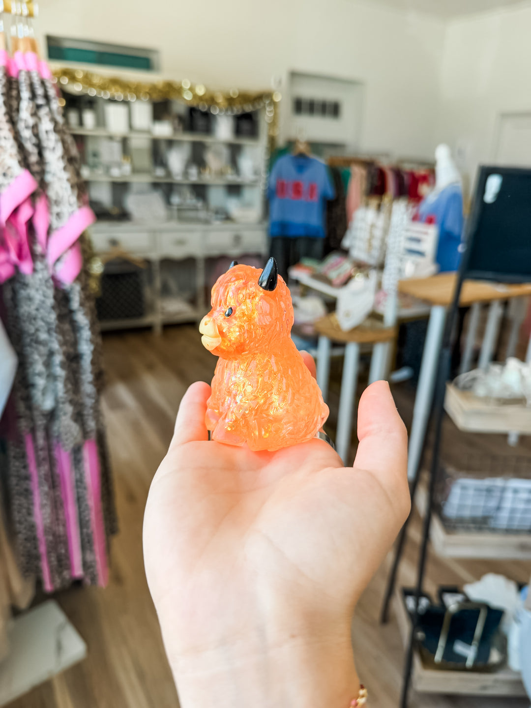 A hand holding a small, translucent orange highland cow squishy with a black horn, set against a blurred boutique background