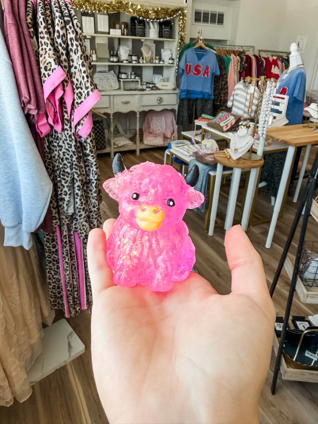 A small, pink highland cow squishy with black horns and a yellow snout is held in a person's hand inside a retail store.