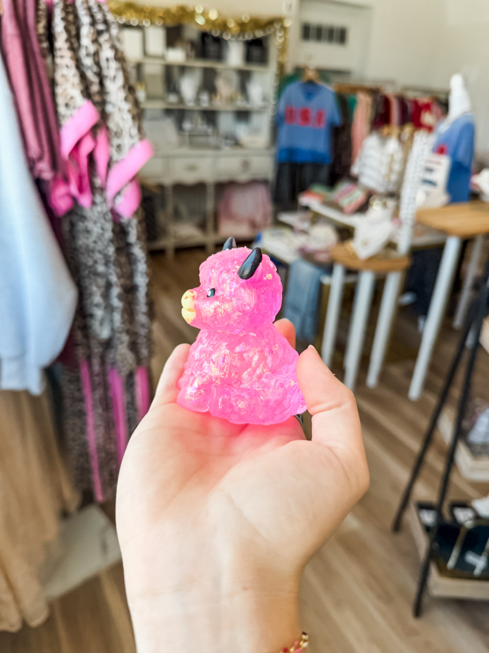 A hand holding a small, glittery pink highland cow squishy toy with black horns and a yellow mouth, set against a blurred boutique background.