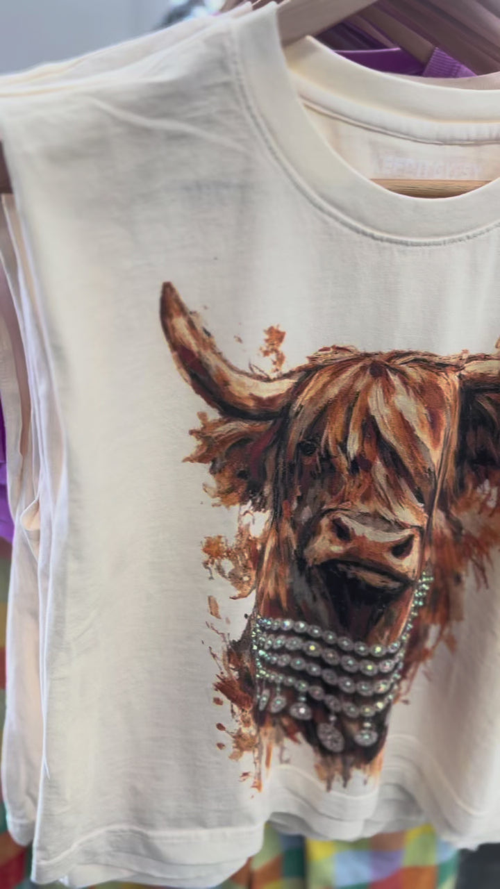 Video showing a cream muscle tank with a highland cow and rhinestone accents hanging on a wooden hanger