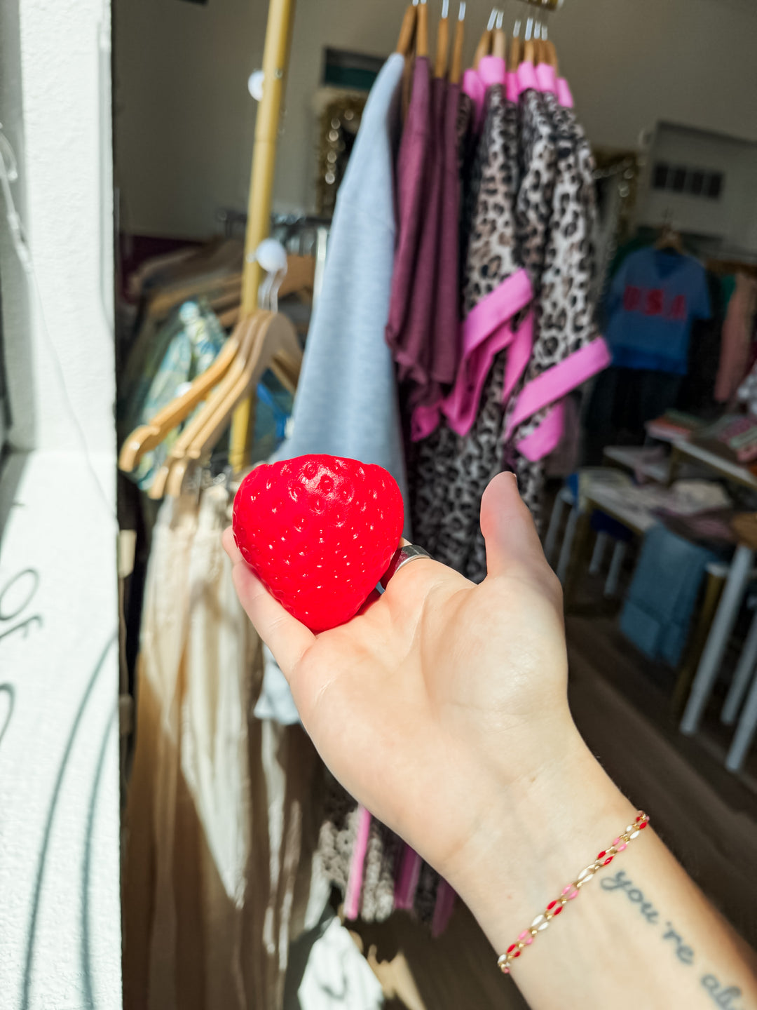 A hand holding a bright red strawberry-squishy sensory toy in front of a clothing rack inside a store.