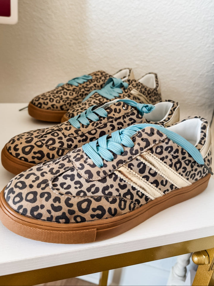 A pair of low-top sneakers with a beige and black leopard print pattern, featuring light blue laces and a gum brown sole, displayed on a white shelf.